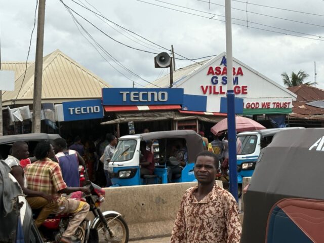 Busy roadside scene outside Asaba Computer Village with buses, traders, and hawkers moving through the market